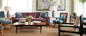 A seat arrangement with a wood and leather coffee table, a series of structural armchairs and a purple sofa. a pair of lamps and vintage art accessorize the space.