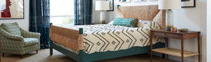 Bedroom scene with a woven rush headboard, zig zag bedcover, and green upholstered lounge chair. Bedside tables & curtains.