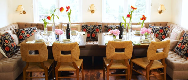 A dining area with a mix of a banquette and Hollywood  at Home's How to Marry a millionaire square back, rush-woven dining chairs. flower on the table. 