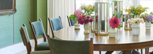 A green dining room with green upholstered dining chairs surrounding an oval table topped with flowers.