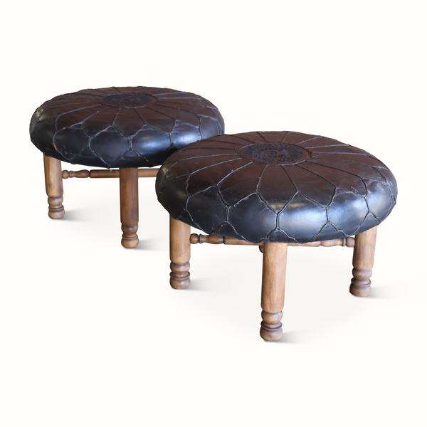 Two round ottomans at an angle, dark leather tops with floral center and wooden feet