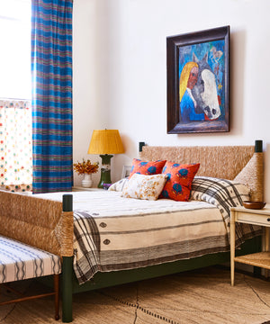 A bed with a rush-woven headboard in a clean room setting with fun pops of blue and orange for art, pillows, and curtains. A lamp and bench and bedside table accompany the focal point of the bed.