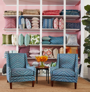 Two upholstered chairs in a  pattern sit in front of a wall unit full of colorful, patterned pillows. The walls are pink.