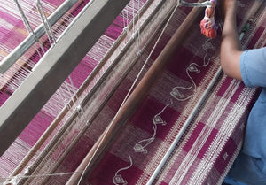 An overhead image of a hand weaver and embroidery working on a fuschia colored textile at a loom.