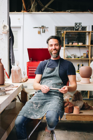 A portrait of ceramicist Natan Moss in his ceramics studio - he's wearing an apron and there are ceramics in the background.