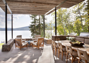 A lakeside outdoor scene under a pergola with a long dining table with chairs and a banquette on the right, and a seating group of four armchairs on the left.