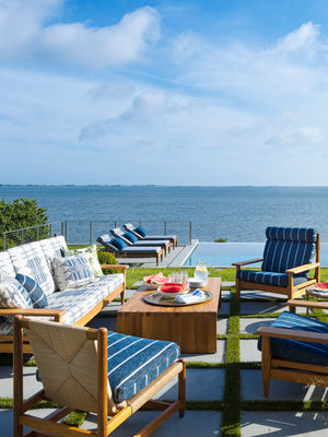 A seaside seating arrangement with teak furniture and blue and white cushions. A platter with watermelon and water cups is on the coffee table.